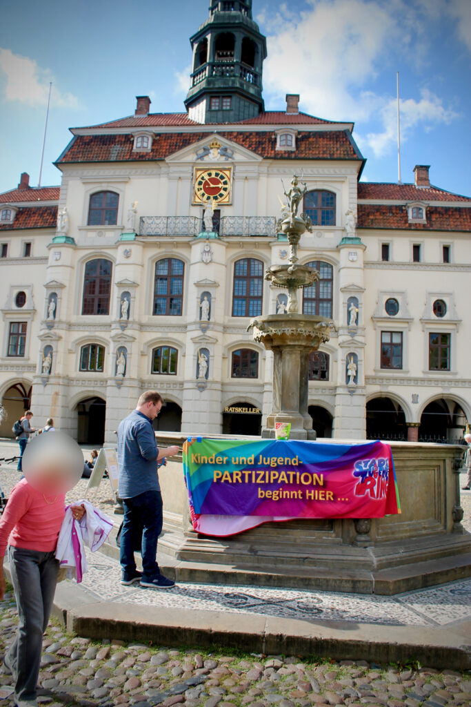 Ein junger Mann hängt ein buntes Banner mit der Aufschrift "Kinder- und Jugendpartizipation beginnt hier... Stadtjugendring Lüneburg" am Luna-Brunnen auf.